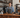 A barista inside a cafe is standing behind the counter, staring at the camera. The cafe is calm and serene.